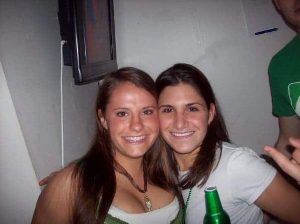 Two girls at a party smiling for a picture. One is holding a beer.