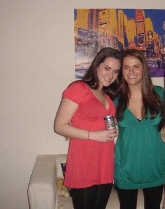 Two girls in an apartment. One wearing a green shirt, one wearing a red shirt and holding a beer