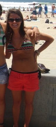 Girl in a bikini top with orange shorts on. Behind her is the beach.