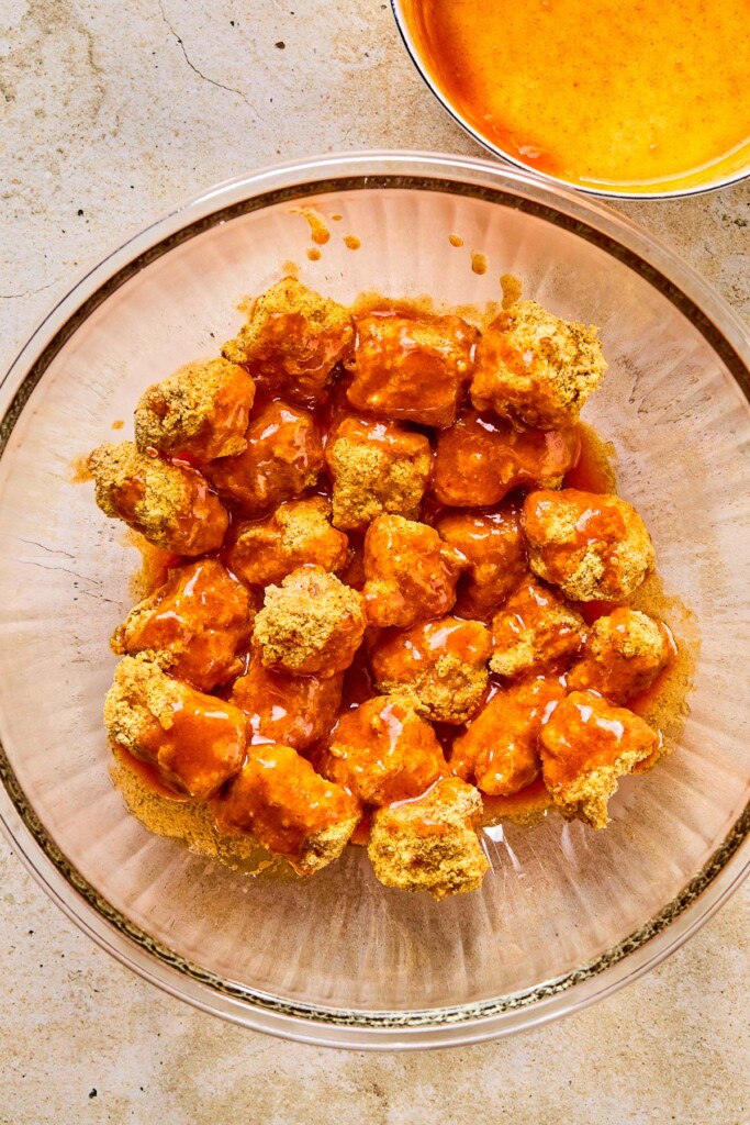 A large bowl of breaded chicken bites with buffalo sauce poured overtop of them. Next to the bowl is the pan the sauce was in.