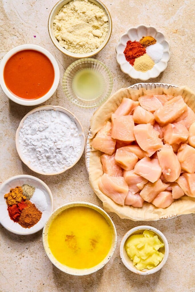 A small bowl of a few piles of different spices, a plate of cubed raw chicken, a small bowl of ghee, a bowl of whisked eggs, another small dish of piles of different spices, a dish of tapioca flour, a small bowl of lemon juice, a small bowl of hot sauce, and a bowl of almond flour on a tan counter.