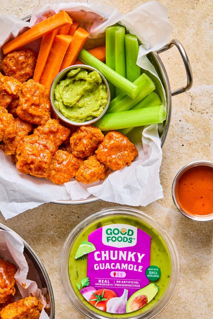 Overhead shot of a large dish of air fryer boneless buffalo wings with carrot sticks, celery sticks, and a small ramekin of guacamole for dipping. Next to the dish is a small ramekin of buffalo sauce, a container of Good Foods Chunky Guacamole, and another dish of wings.