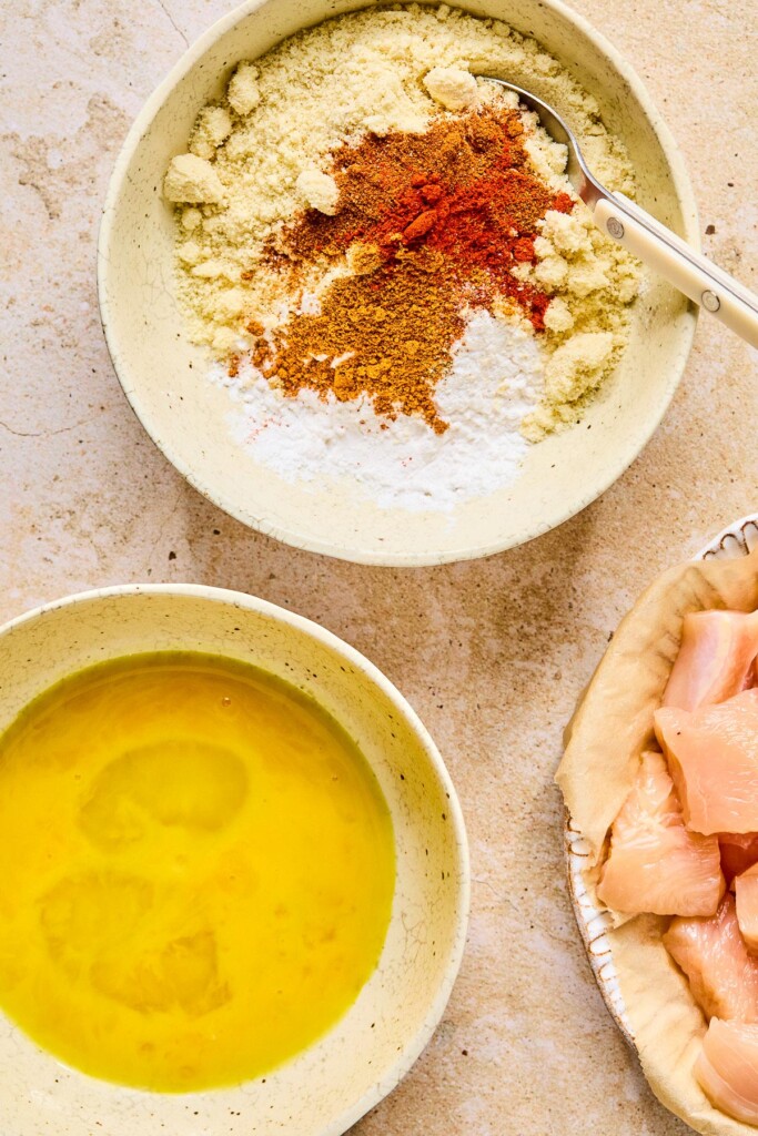 Overheat shot of a bowl of two kinds of flours and spices with a whisk in it before it is mixed together. Next to it is a plate of raw cubed chicken and a bowl with bowl with whisked eggs in it.