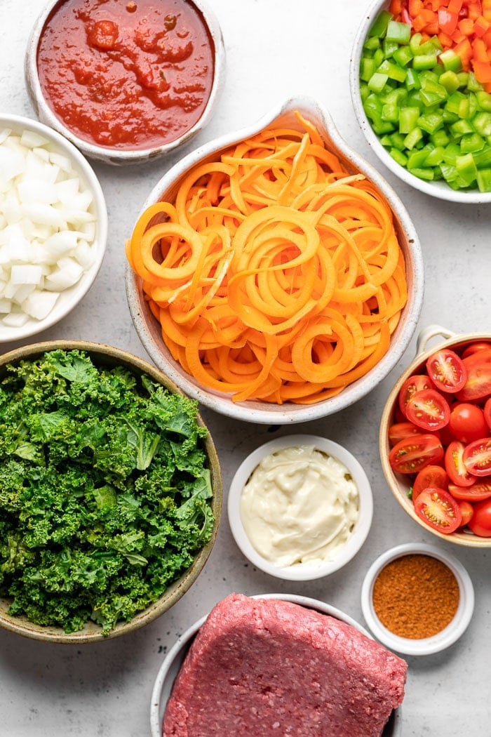 Overhead shot of a bowl with salsa, bowl with chopped peppers, bowl of sweet potato noodles, bowl of diced tomatoes, bowl of raw ground beef, bowl of chopped kale, bowl of diced onion, bowl of mayo, and bowl of taco seasoning.