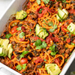 Close up of a large pan filled with ground beef taco casserole topped with cilantro and avocado. Behind it is a small bowl of diced tomatoes.