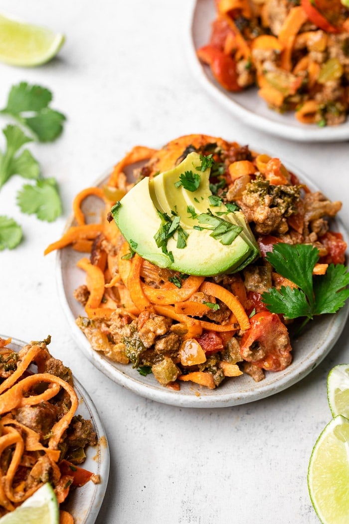 Plate of easy taco casserole topped with sliced avocado and cilantro. Next to it is two more plates, cilantro, and lime wedges.