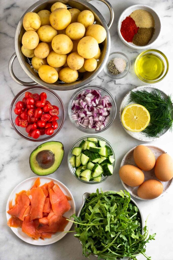 Marble counter top with a colander of baby potatoes, a bowl of spices, a bowl of salt and pepper, a bowl of olive oil, a plate of fresh herbs and a half a lemon, a plate of 4 eggs, a bowl of arugula, a plate of smoked salmon, a bowl of diced cucumber, half an avocado, a bowl of diced tomatoes, and a bowl of diced onions.