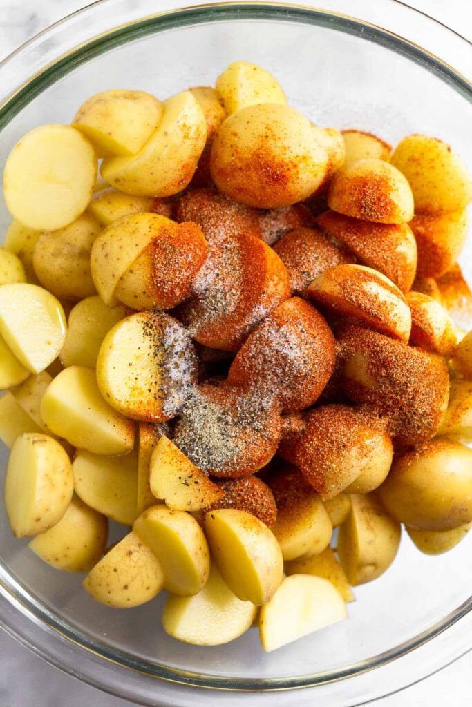 A large glass dish filled with diced potatoes with oil and spices poured overtop before they are mixed.