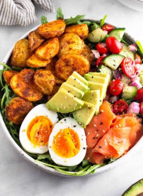 Breakfast bowl filled with greens, roasted potatoes, tomato and cucumber salad, smoked salmon, sliced avocado, and a hard-boiled egg and topped with everything but the bagel seasoning. Next to it is half an avocado.