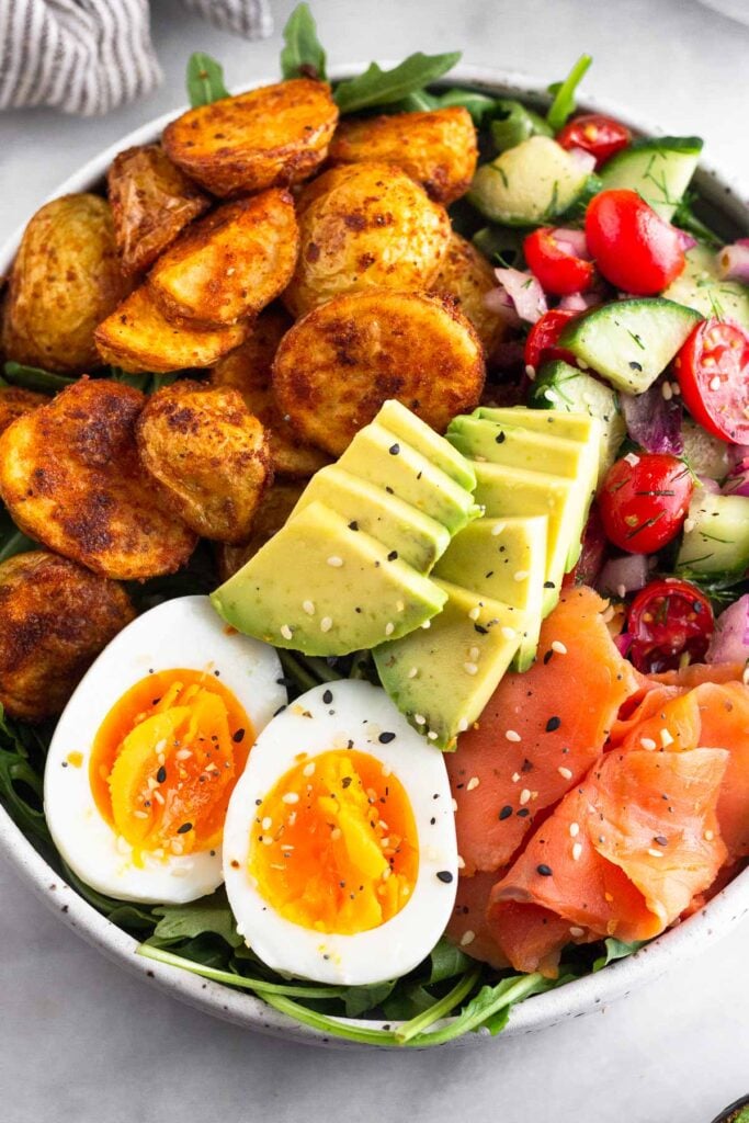 Close up of a breakfast bowl with greens, roasted potatoes, tomato and cucumber salad, smoked salmon, sliced avocado, and a hard-boiled egg. It is sprinkled with everything but the bagel seasoning.