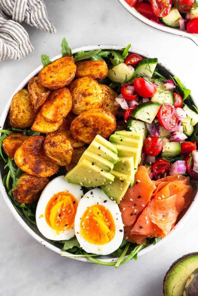 A bowl filled with greens, roasted potatoes, tomato and cucumber salad, smoked salmon, sliced avocado, and a hard-boiled egg. It is topped with everything but the bagel seasoning. Around it is half an avocado, a bowl of tomato and cucumber salad, and a kitchen towel.