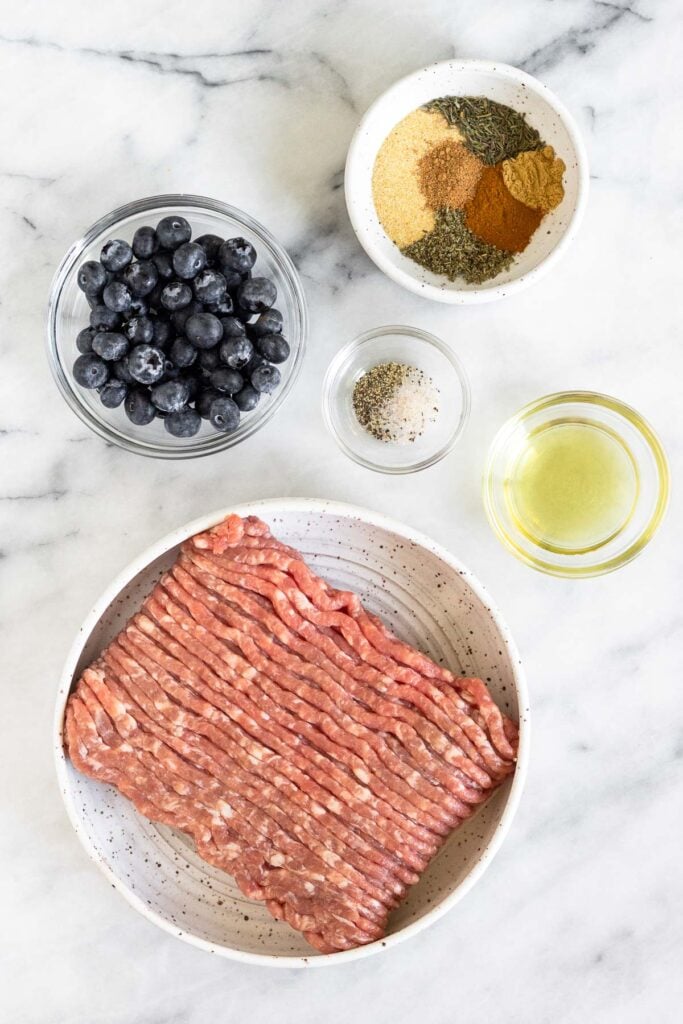 White marble counter with a bowl of spices, a bowl of oil, a bowl of ground pork, a bowl of salt and pepper, and a bowl of blueberries.