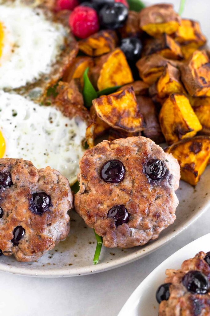 A close up of a plate with breakfast sausage patties, roasted sweet potatoes, berries, and fried eggs. Off to the side is a plate of more sausage patties.