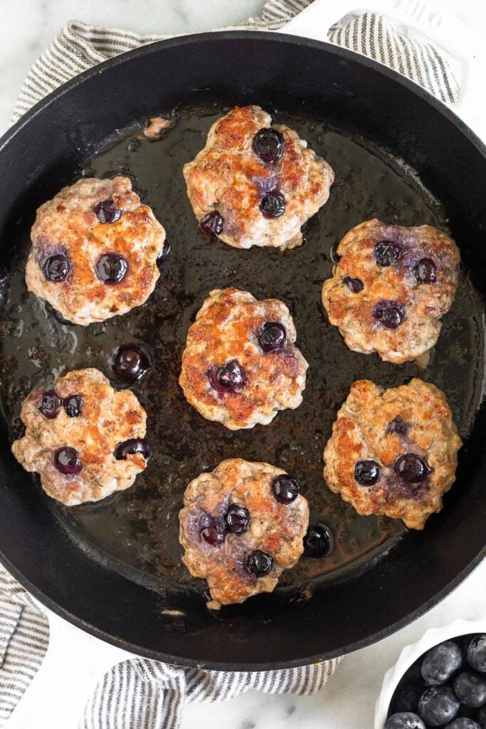 A pan filled with blueberry breakfast sausage sitting on a striped kitchen towel. Next to it is a small dish of fresh blueberries.