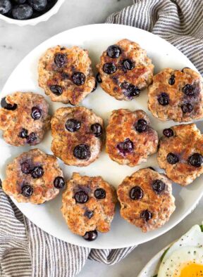 A white plate of blueberry breakfast sausage patties. They plate is sitting on a kitchen towel and next to it is a bowl of blueberries.