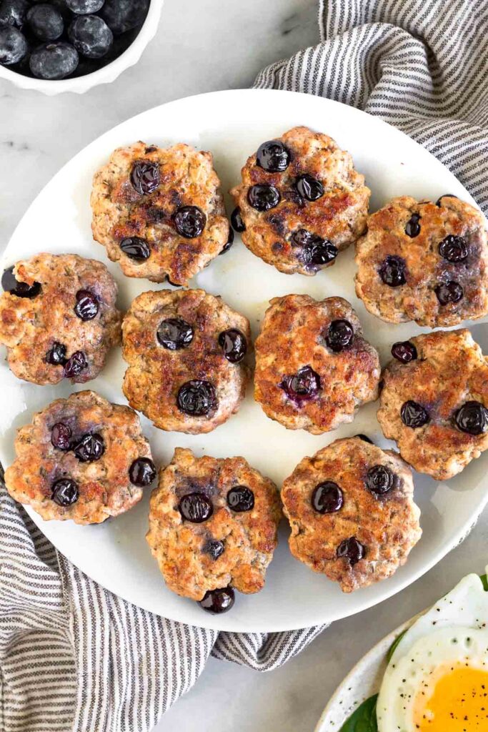A white plate of blueberry breakfast sausage patties. They plate is sitting on a kitchen towel and next to it is a bowl of blueberries.