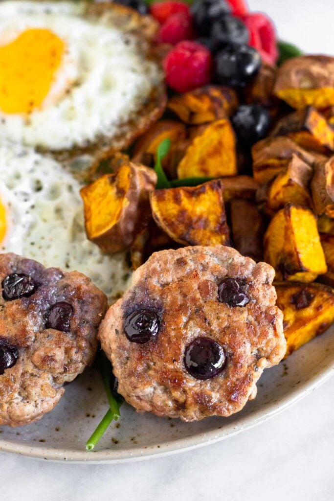 A plate of two homemade breakfast sausages, fried eggs, berries, and roasted sweet potatoes on greens.