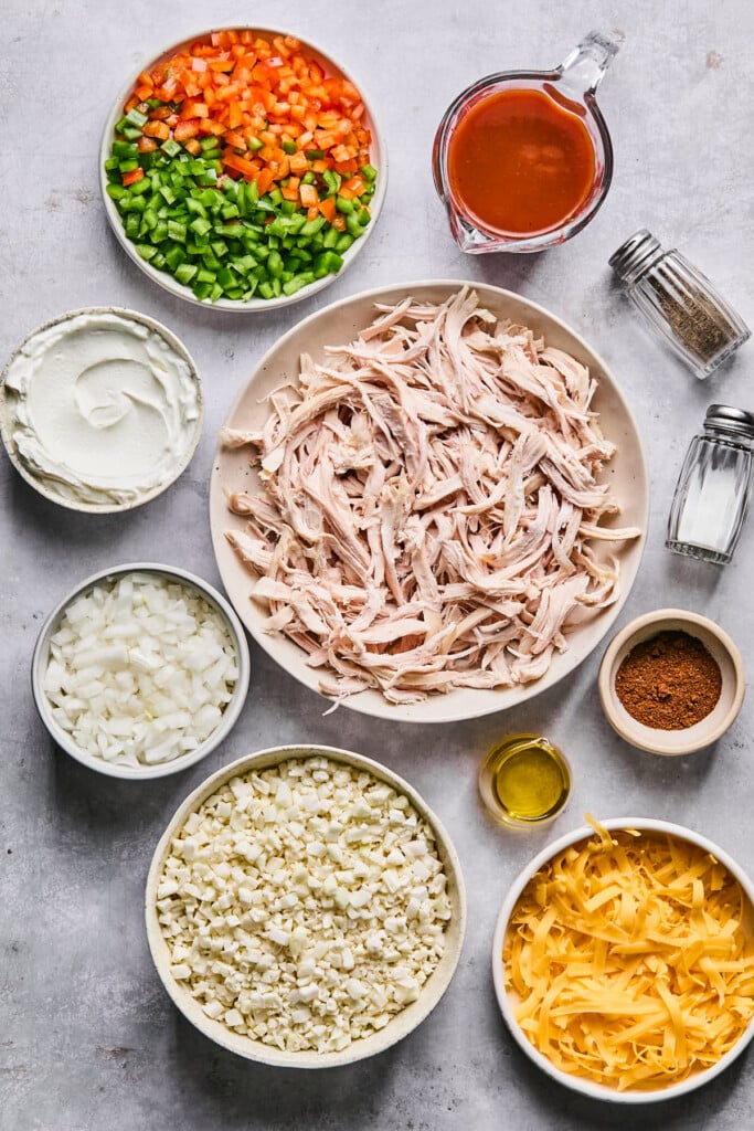 Grey concrete counter with a measuring cup of enchilada sauce, a bowl of shredded chicken, a salt and pepper shaker, a small dish of spices, a small dish of olive oil, a bowl of shredded cheese, a bowl of uncooked cauliflower rice, a bowl of diced onions, a bowl of greek yogurt, and a bowl of diced red and green bell peppers.