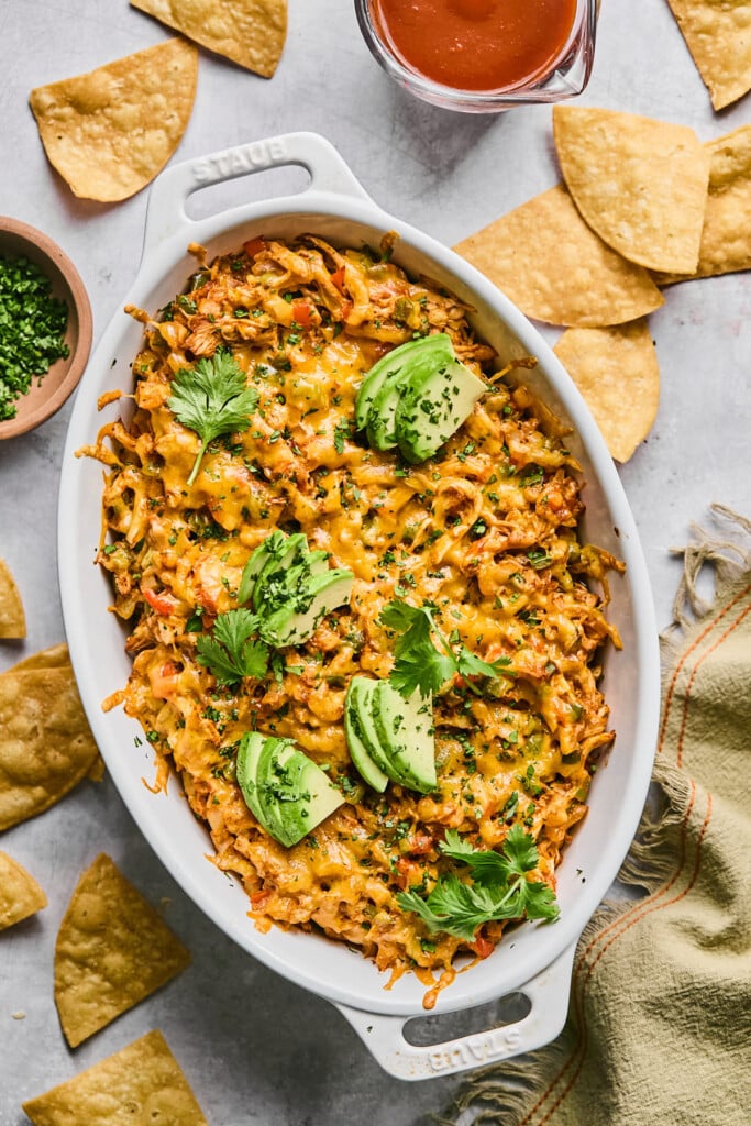 Cheesy chicken enchilada dip in a white dish topped with sliced avocado and cilantro. Around the pan are some tortilla chips, a bowl of cilantro, a mixing cup of enchilada sauce, and a tan linen.