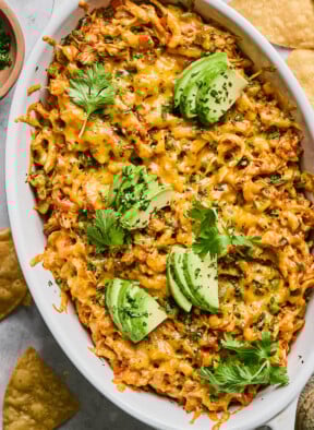 Au gratin dish filled with chicken enchilada dip topped with melted cheese and garnished with cilantro and sliced avocado. Around the dish are some tortilla chips.