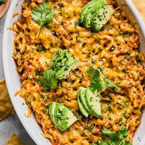 Au gratin dish filled with chicken enchilada dip topped with melted cheese and garnished with cilantro and sliced avocado. Around the dish are some tortilla chips.