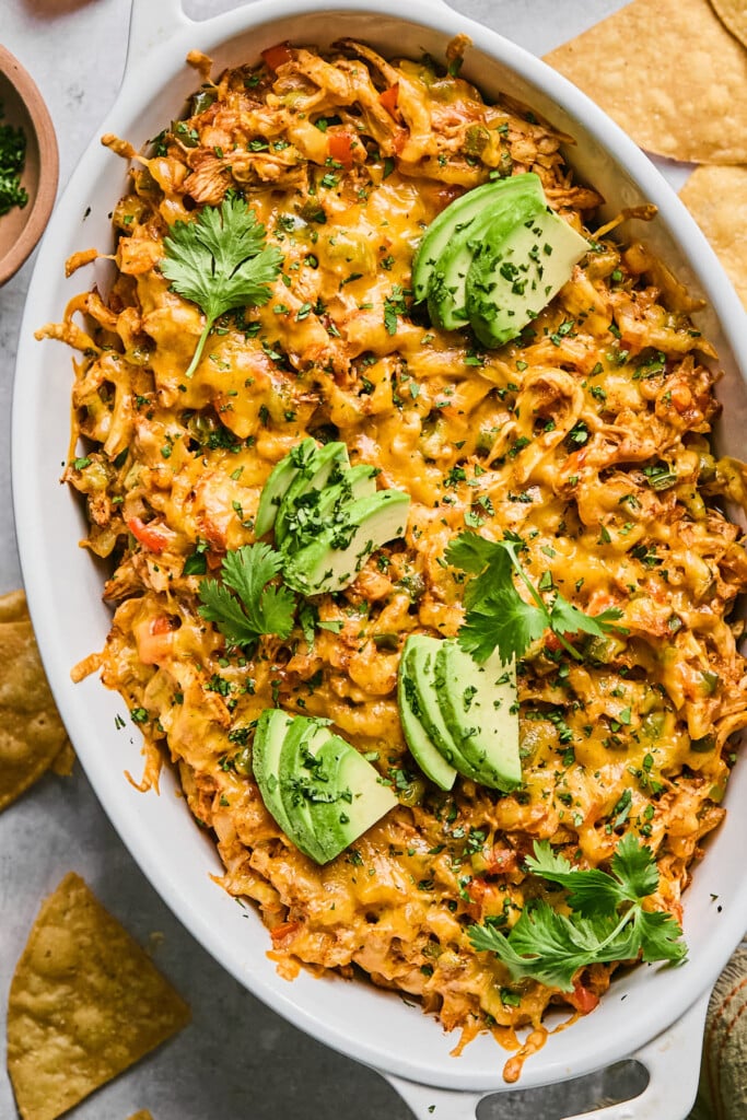 Au gratin dish filled with chicken enchilada dip topped with melted cheese and garnished with cilantro and sliced avocado. Around the dish are some tortilla chips.