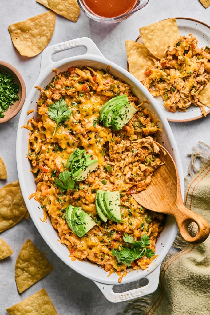 White baking dish filled with chicken enchilada dip topped with cilantro and sliced avocado. A large wooden spoon is also in the dish. Around the dish are some tortilla chips, a bowl of cilantro, a cup of enchilada sauce, a tan linen, and a small plate with some dip and chips on it.