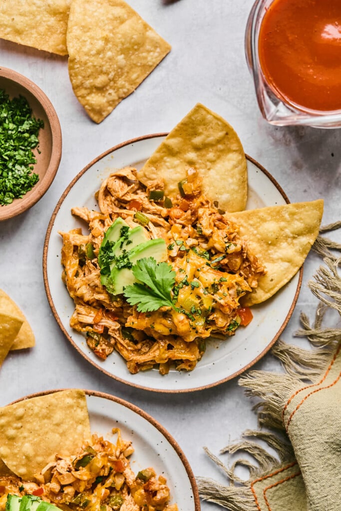 Chicken enchilada dip on a plate garnished with sliced avocado and cilantro. Also on the plate are some tortilla chips. Around the plate is a bowl of chopped cilantro, tortilla chips, a measuring cup of enchilada sauce, a tan linen, and another plate of dip and chips.