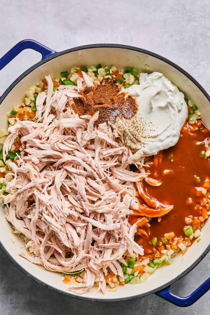Large enameled pan filled with diced sautéed veggies, shredded chicken, spices, greek yogurt, and enchilada sauce before it is mixed together.