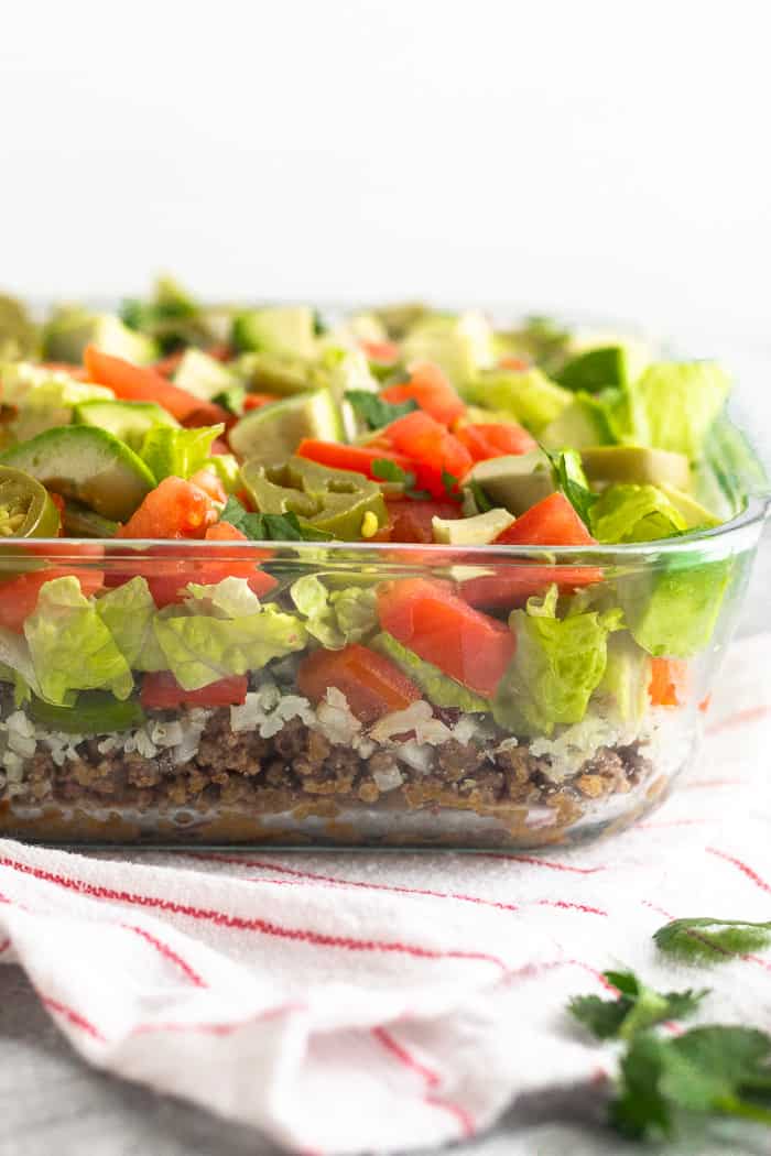 Straight on view of Whole30 and paleo 7 layer dip. It is in a glass container with a layer of beef, cauliflower rice, peppers and onions, lettuce, tomato, avocado, and jalapeños.
