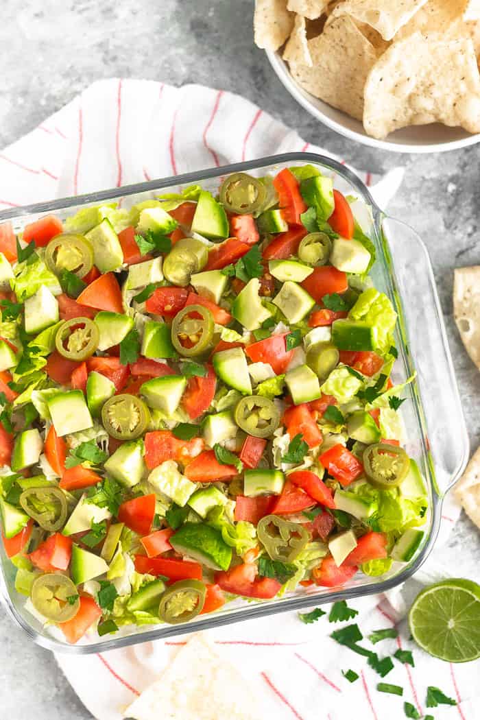 Overhead shot of paleo 7 layer dip in a large glass container. It is sitting on a red and white stripped towel with a bowl of chips, half a lime, and cilantro around it.