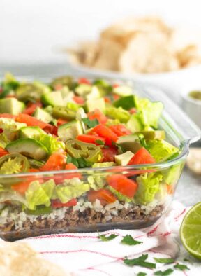 Glass baking dish filled with paleo 7 layer dip. The dish is sitting on a red and white stripped towel with chips, cilantro, and half a lime around it. Behind it is a bowl of chips.