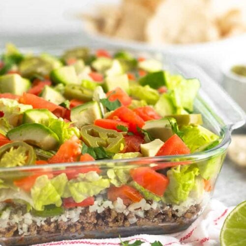Glass baking dish filled with paleo 7 layer dip. The dish is sitting on a red and white stripped towel with chips, cilantro, and half a lime around it. Behind it is a bowl of chips.