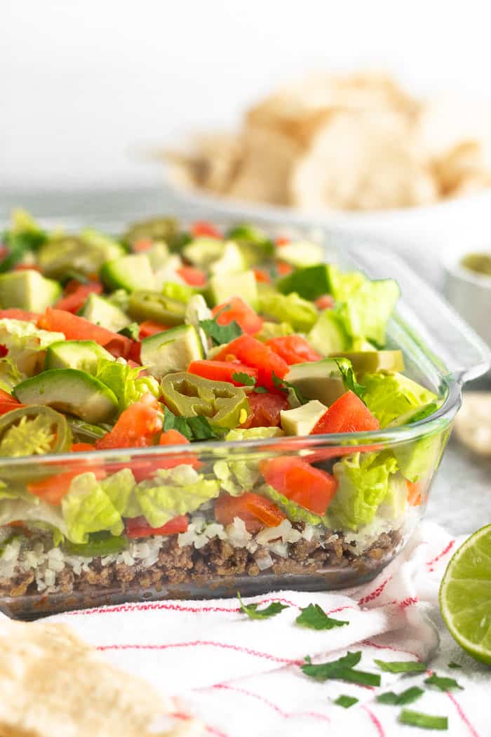 Glass baking dish filled with paleo 7 layer dip. The dish is sitting on a red and white stripped towel with chips, cilantro, and half a lime around it. Behind it is a bowl of chips.