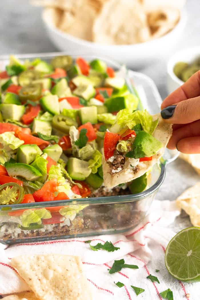 Large glass dish filled with paleo 7 layer dip. A chip is in front of the dish with a dip on the chip as it was just scooped out.