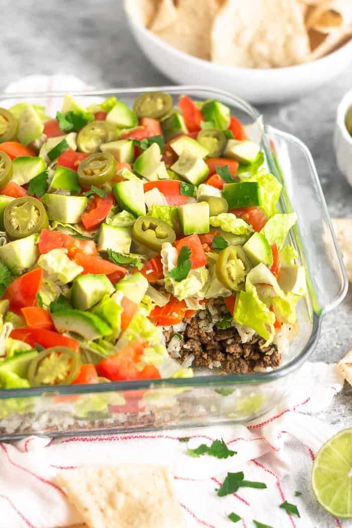 A glass dish filled with low carb paleo 7 layer dip with a scoop taken out of it. It is surrounded by a white and red stripped dish towel, a bowl of chips, half a lime, and cilantro.