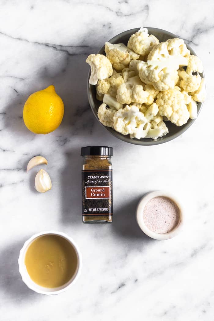 A bowl of cauliflower florets, salt, small bowl of tahini, cumin, 2 garlic cloves, and a lemon on a white marble countertop.