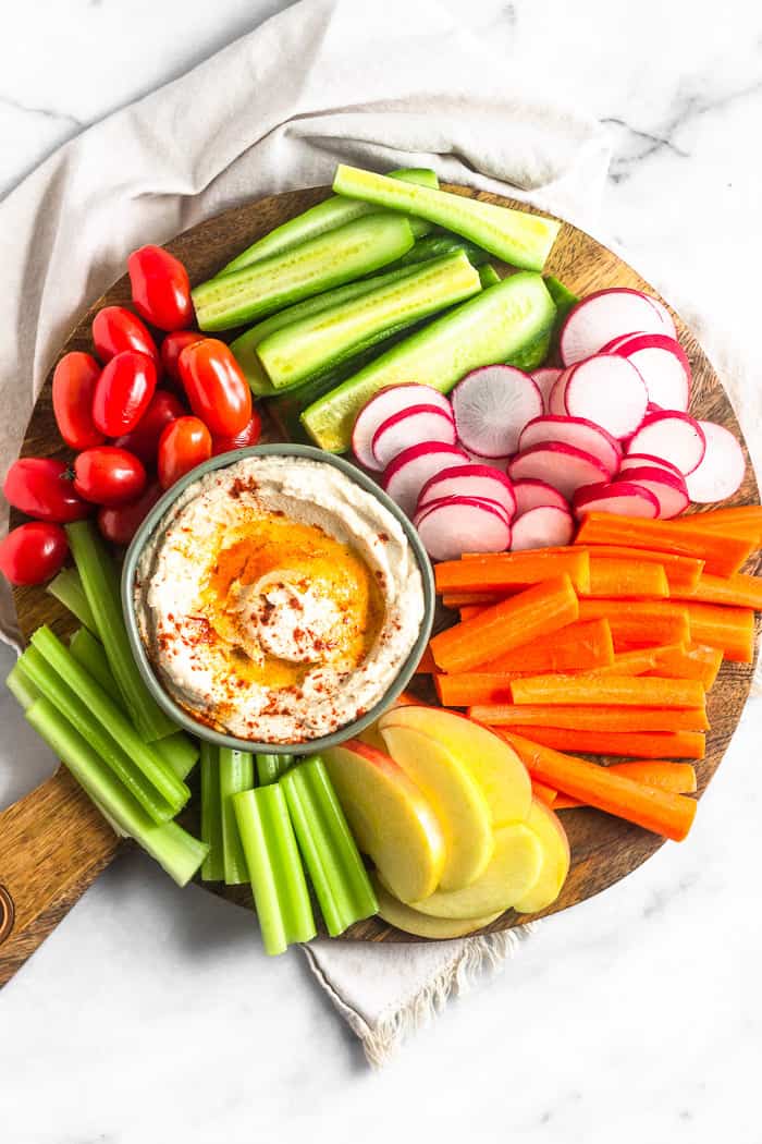 Large wooden platter filled with fresh cut veggies and fruit. In the middle is a bowl of paleo cauliflower hummus drizzled with olive oil and paprika.