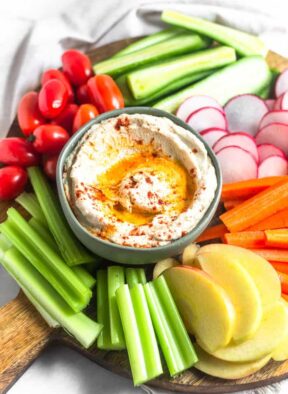 A circular wooden cutting board filled with a bowl of roasted cauliflower hummus surrounded by a bunch of fresh cut fruits and veggies.