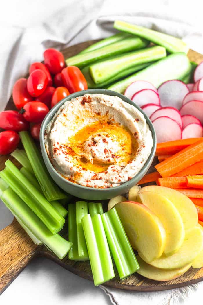 A circular wooden cutting board filled with a bowl of roasted cauliflower hummus surrounded by a bunch of fresh cut fruits and veggies.