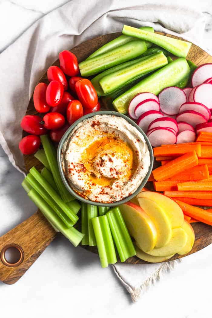 Overhead shot of a wooden serving dish on a white linen. On the serving dish is a bowl of pale cauliflower hummus surrounded by fresh cut veggies and fruit.