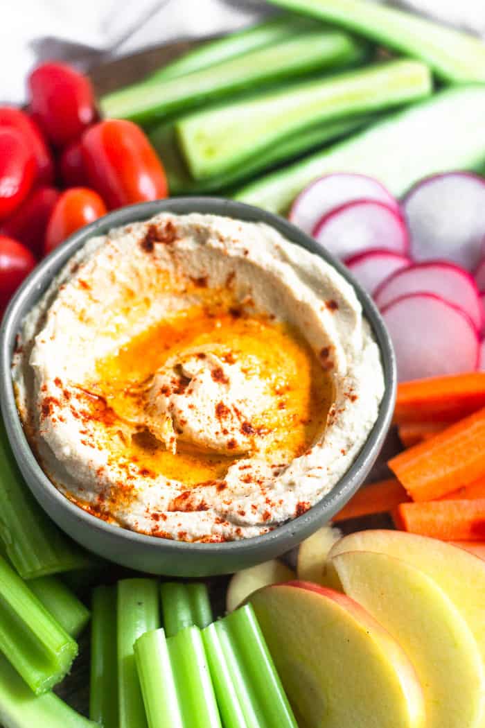 Close up of a bowl of roasted cauliflower hummus with olive oil and paprika on top of it. It is surrounded by fresh veggies and fruits.