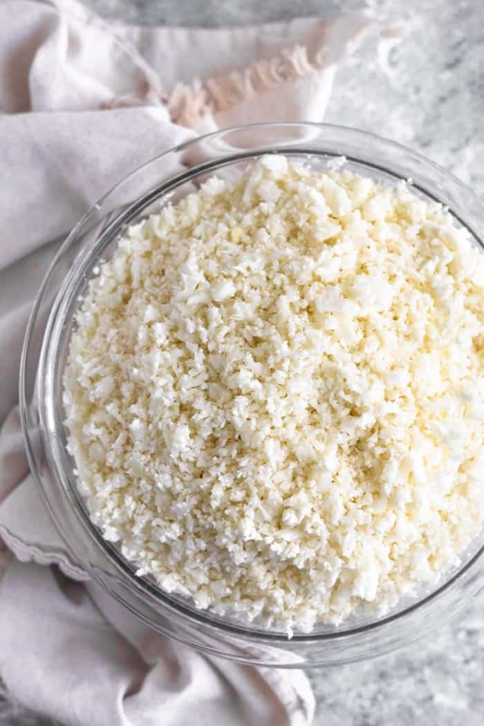 A large glass bowl filled with fresh cauliflower rice. The bowl is sitting on a tan linen.
