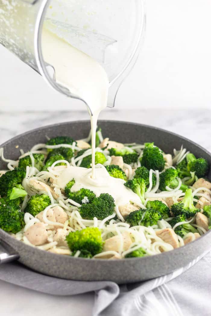 Large grey skillet filled with Whole30 chicken and broccoli. A blending is pouring dairy free Alfredo sauce over it.