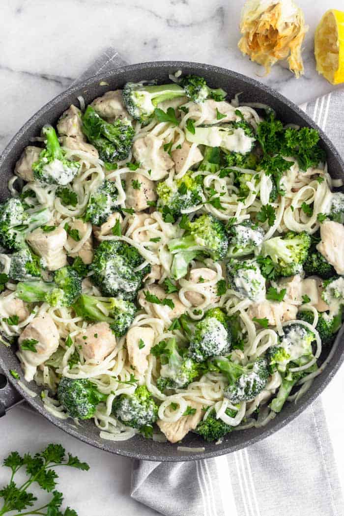 Large skillet filled with Whole30 Chicken and Broccoli Alfredo. It is sitting on a grey towel and next to it is some fresh parsley, a head roasted garlic, and half a lemon.
