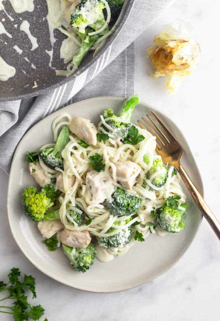 Overhead shot of a white plate with Whole30 Chicken and Broccoli Alfredo on it. A gold fork is resting on the plate as well. It is next to a large skillet, a head of roasted garlic, and some parsley.