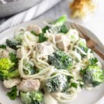 A white plate with Whole30 Chicken and Broccoli Alfredo with a gold fork resting on the plate. Behind it is a head of roasted garlic and the front of a silver skillet.