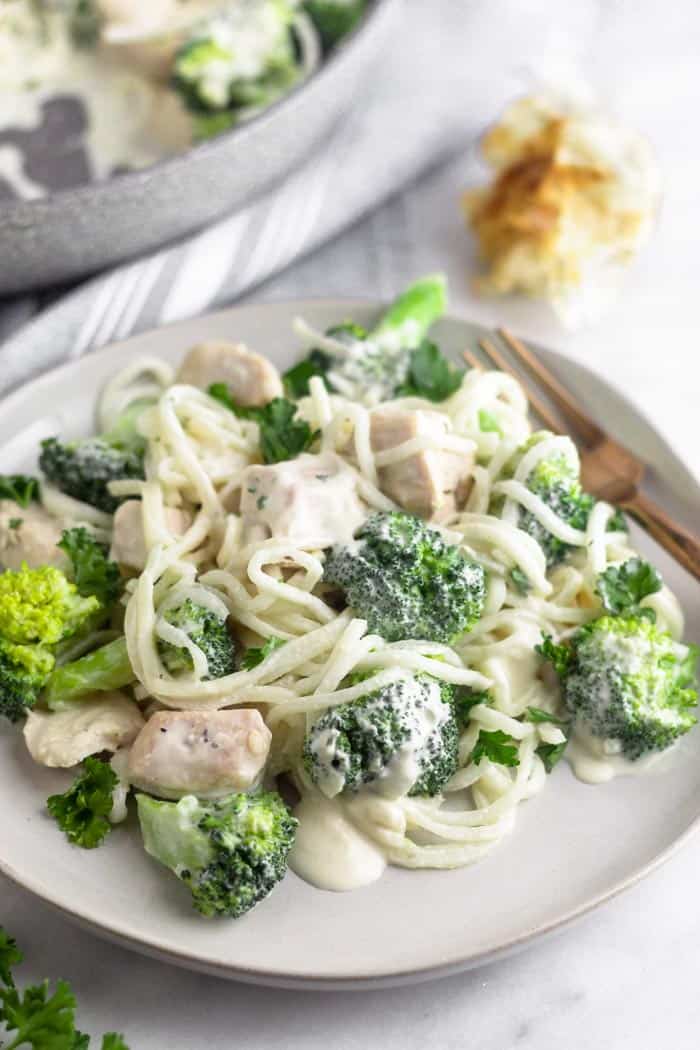 A white plate with Whole30 Chicken and Broccoli Alfredo with a gold fork resting on the plate. Behind it is a head of roasted garlic and the front of a silver skillet.