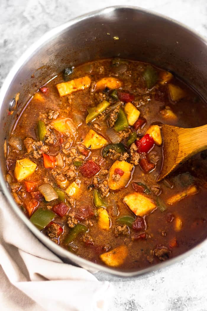 Large pot filled with paleo beef and plantain chili sitting on a tan linen. A wooden spoon is sticking out of the pot.