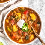 White bowl filled with Whole30 beef and plantain chili topped with coconut yogurt and sliced jalapeños. A gold spoon is sticking out of the bowl. Around the bowl is a tan linen, another bowl of chili, and a plate with lime wedges.